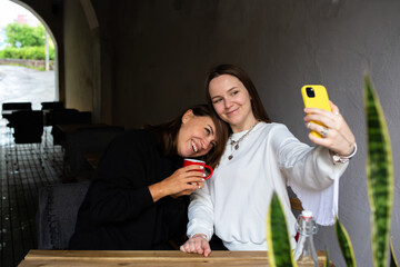 Two women friends have fun outdoors in cafe and take a selfie on a smartphone