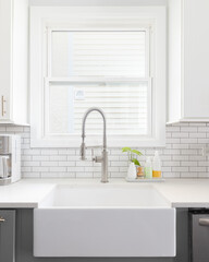 Fototapeta na wymiar A kitchen sink detail shot with white and grey cabinets, a farmhouse sink, and white subway tile backsplash.