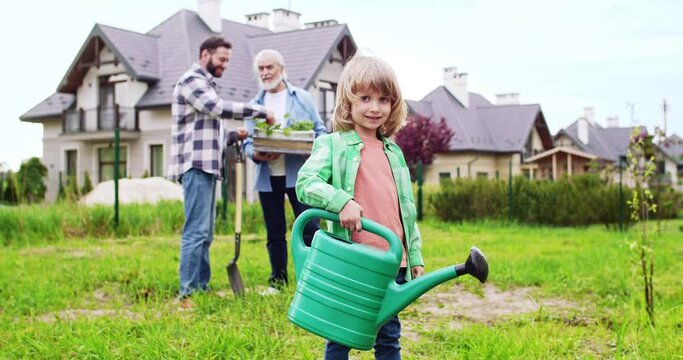 Portrait of cute Caucasian happy little boy with watering pot in hands smiling to camera. Father and grandfather talking on background. Planting trees and plants in garden at summerhouse. Outside.
