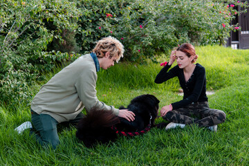 Youthful couple are petting their black dog while resting on the grass