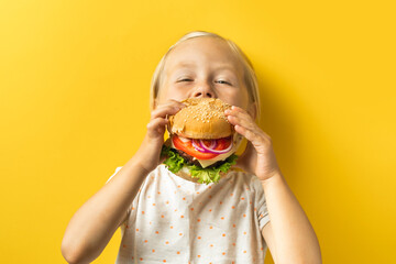 Cute little caucasian girl with blonde hair enjoying burger on a yellow background. Happy kid...