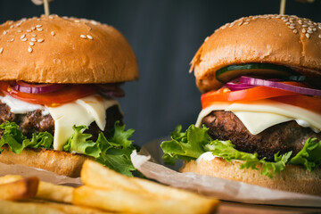 Big beef patty burgers with cheese tomato purple onion cucumber and lettuce next to french fries on wooden black stand on black background