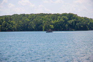Fototapeta na wymiar a motor boat sailing across the rippling blue waters of Lake Allatoona surrounded by lush green trees with blue sky and clouds at Victoria Beach in Acworth Georgia USA