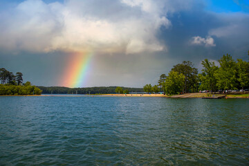 a gorgeous rainbow over Lake Allatoona with a beach and rippling blue lake water surrounded by lush green trees, grass and plants with clouds at Victoria Beach in Acworth Georgia USA