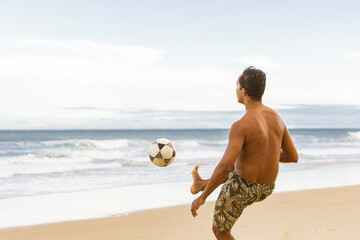 Back shot of Latin male playing football soccer at the beach on vacation, kicking the ball. Copy...