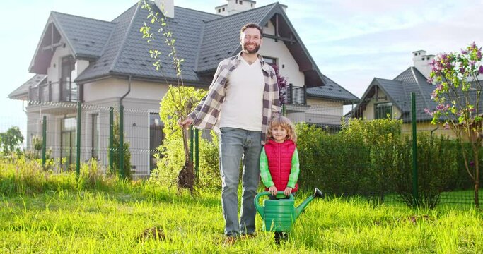 Portrait of happy young Caucasian father with small tree with root in hand and little cute son with watering pot. Garden work in summer. Planting trees concept.