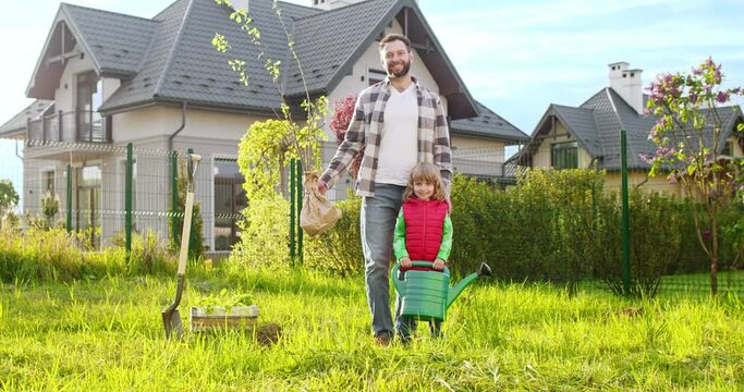 Portrait shot of cheerful young Caucasian father with small tree in hand and little cute son with watering pot. Orchard work in summer. Planting trees concept. Outdoor.
