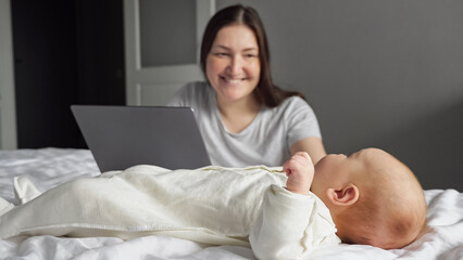 Mother freelancer looks after baby girl while working on laptop in bedroom. Newborn child wiggles...