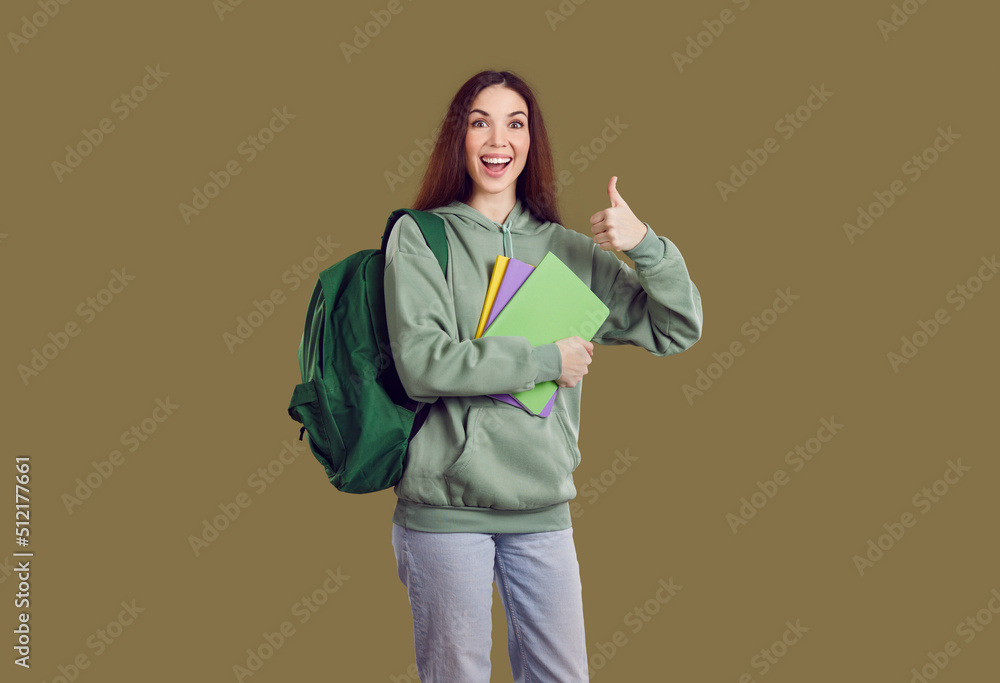 Wall mural Happy cheerful beautiful college or university student girl in green hoodie with backpack standing isolated on solid greenish brown color background, holding her books, showing thumbs up and smiling