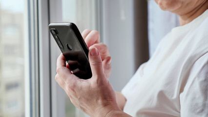 Old lady in white t-shirt uses modern smartphone. Woman with short haircut scrolls web pages on black smartphone standing near window