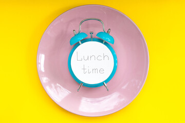 Handwritten text "lunch time" On blue alarm clock on the pink plate. Yellow background. Concept of time to break. 