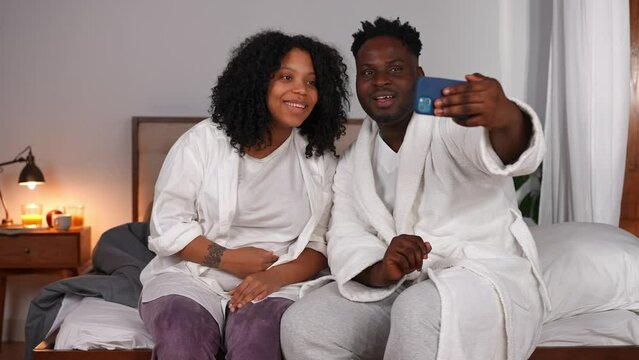 Positive African American man and pregnant woman waving talking at smartphone web chat. Smiling happy relaxed couple messaging online sitting on bed at home indoors. Convenience and domestic lifestyle