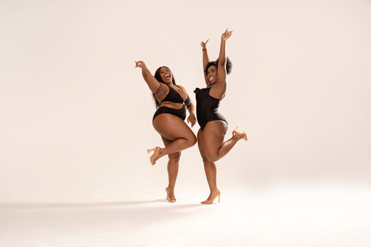 Full photo of superb plus size dark skinned two women in black fashionable swimwears, laughing and dancing, having fun together. Concept of body acceptance, body positivity