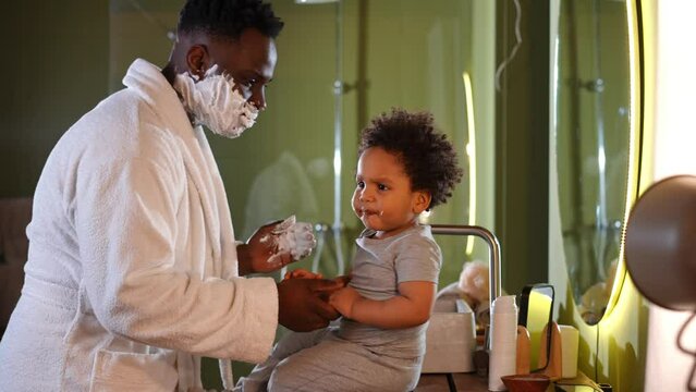 Side view cute little African American boy sitting in bathroom with father shaving playing with child. Curios charming kid enjoying morning with young handsome man in white shave foam
