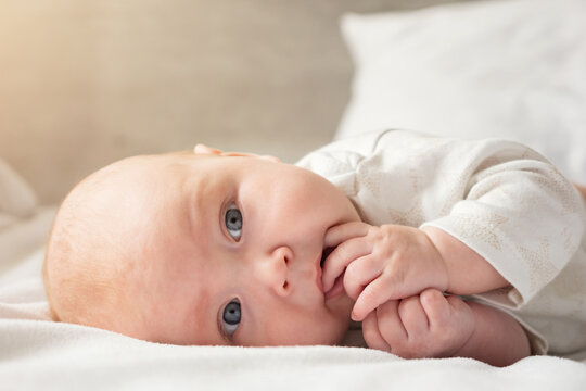 Baby girl lies on side on big white parent bed. Newborn child with grey eyes and plump cheeks lies in bedroom putting his fingers in his mouth and sucking his fingers, sunlight