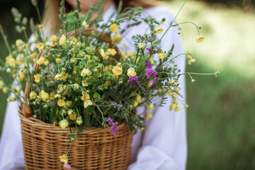 A girl in a white blouse holds a wicker basket with a bouquet of wild flowers. Summer walk in the field. midsection