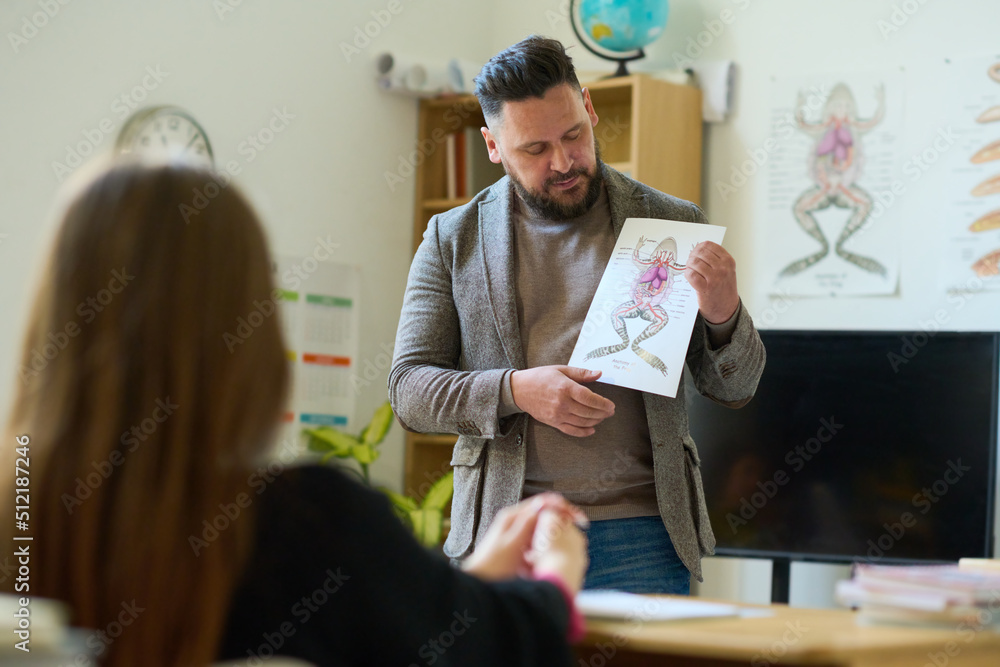 Poster confident mature biology teacher showing paper with frog picture to student while making presentatio