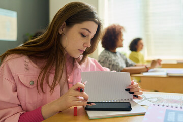 Teenage girl in pink denim jacket texting in smartphone at lesson behind page of copybook while sitting by desk against her classmates