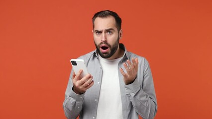 Sad irritated angry young brunet man 20s years old wears blue shirt using mobile cell phone swear hears fake news unexpected rumor has some problems isolated on plain orange background studio portrait