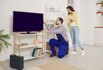 Professional workman in workwear installing large TV monitor in house of female client. Male cable...