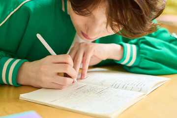 Teenage girl in green jacket bending over desk while making notes in copybook or writing down lecture after teacher at lesson