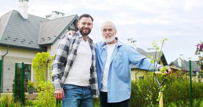 Portrait shot of happy old Caucasian father at small just planted tree and handsome adult young son smiling together to camera. Garden work in summer. Planting trees concept.