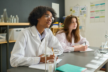 Two clever teenage students in labcoats looking at hteir teacher while sitting by desks at lesson...