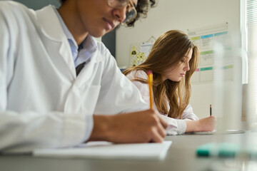 Youthful girl in labcoat making notes in copybook during laboratory work at lesson of chemistry while sitting next to her classmate