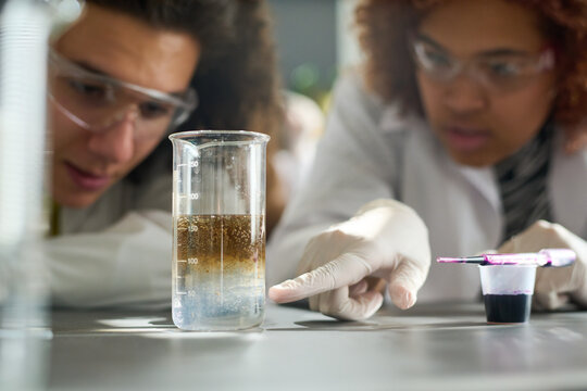 Teenage student in protective gloves pointing at glass containing chemical dissolver during experiment at lesson of chemistry
