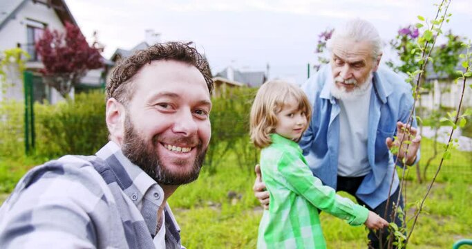 POV of happy Caucasian son taking video and showing small cute son with old grandfather in garden at little tree. Videochat call. Waving with hands. Videochatting.