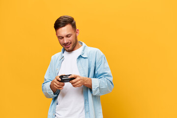 Overjoyed tanned handsome gamer man in blue basic t-shirt hold joystick look at gamepad posing isolated on orange yellow studio background. Copy space Banner Mockup. People lifestyle concept - obrazy, fototapety, plakaty