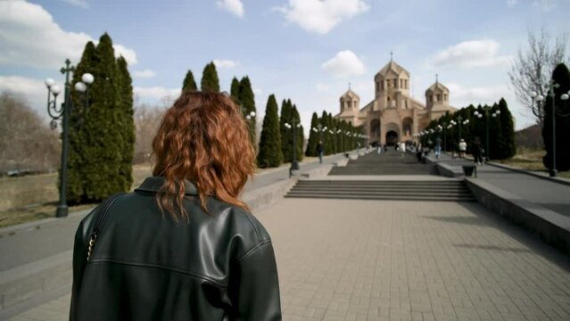 Travelling woman walking towards Saint Gregory the Illuminator Cathedral. Action. Yerevan Cathedral and stairs decorated by growing tujha trees on blue cloudy sky background.