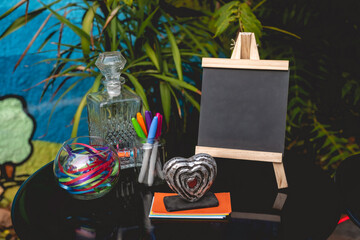 Round black table for messages with little blackboard, colored pens, colored papers, ribbons, glass bottle and little heart decoration with plants background in a latin wedding in the garden