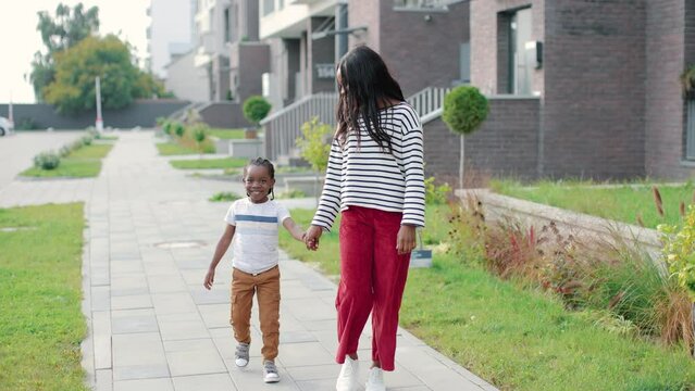 Happy positive young African American woman walking with little cute son outdoor on street. Young beautiful mother with joyful small child boy walk in the city and smiling. Family together mom and kid