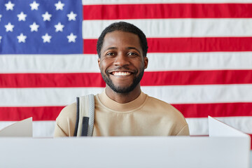 Portrait of smiling black man standing in voting booth and smiling at camera against American flag