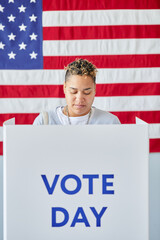 Vertical portrait of young black woman standing in voting booth on election day against American flag