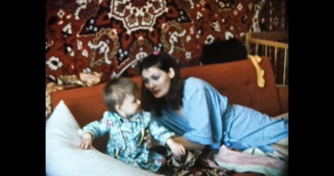 Mother feeds baby with spoon in home room. Young, beautiful woman begs her child to eat in Soviet apartment interior. Happy parents care about child. Vintage color film. Family archive. Retro 1980s