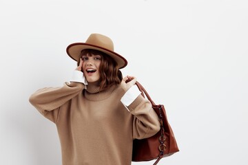 a sweet, happy woman stands on a white background in a beige autumn sweater with a hat on her head and holding a bag on her shoulder, smiling happily with her hand near her face