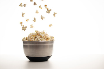 close-up of an aluminium bowl filled with popcorn and falling or floating popcorn on a white background and grey table or worktop. 