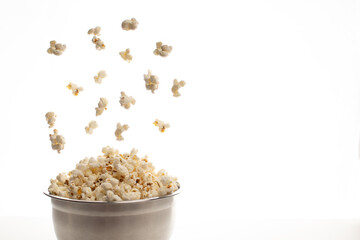 close-up of an aluminium bowl filled with popcorn and falling or floating popcorn on a white background and grey table or worktop. 