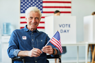 Front view portrait of smiling senior man with disability holding USA flag in voting station, copy...
