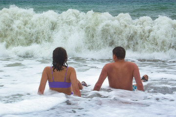 Rear view of a young couple sitting on the shore in sea water. There is sea foam around the girl...