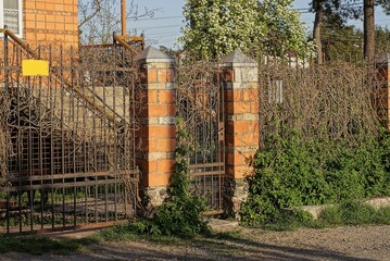 an iron door and part of a fence made of brown bricks and metal rods in a forged pattern overgrown...