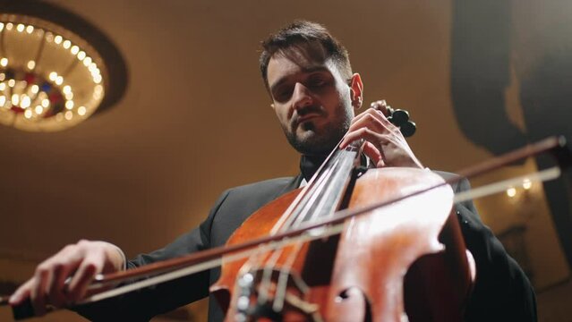 inspired male musician is playing cello on scene of old opera house, portrait of cellist in music hall