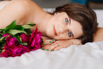 Obraz na płótnie Canvas a gentle brunette woman in bed with a bouquet of peonies. morning of holiday.