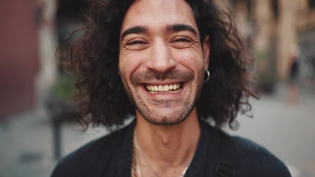 Close-up of young attractive italian guy with long curly hair and stubble looks into the camera and smiles at old buildings background. Stylish man with an earring in his ear and lot of chains