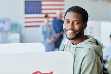 Side view portrait of young black man voting in booth on election day and smiling at camera, copy...