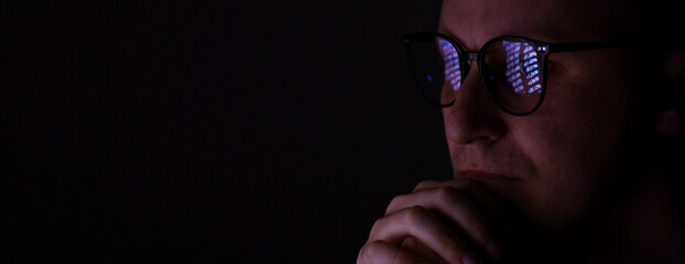 male working online late. Close up side view of focused man wears computer glasses for reducing eye...