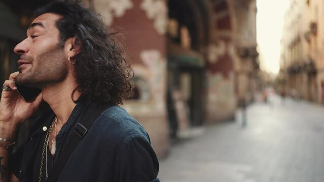Young attractive italian guy with long curly hair and stubble is using mobile phone at old buildings background. Stylish man with an earring in his ear and lot of chains talking on smartphone
