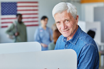 Side view portrait of handsome senior man voting in booth on election day and smiling at camera,...